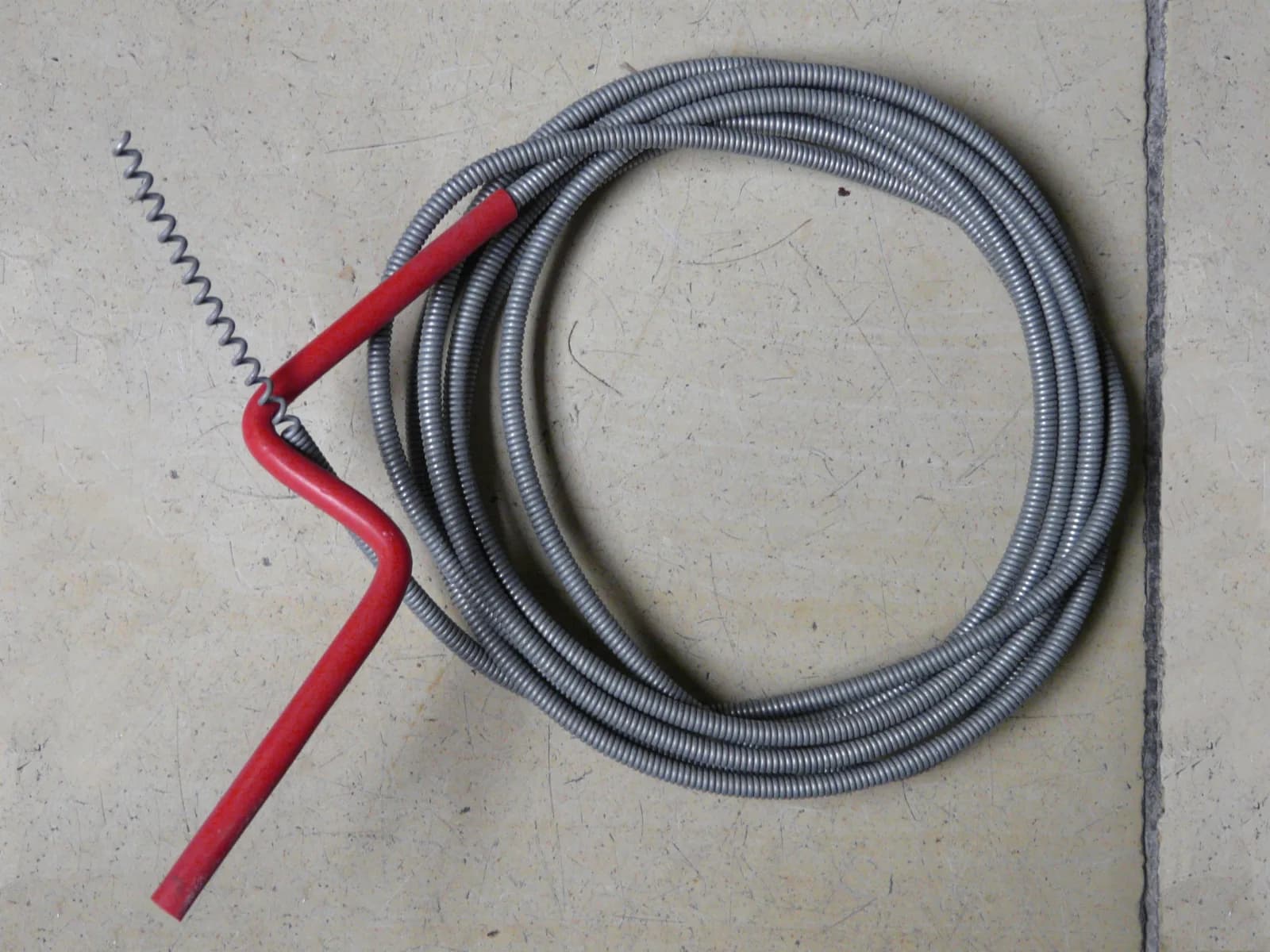 Hand-crank drain snake with coiled steel cable and red handle on a tiled floor