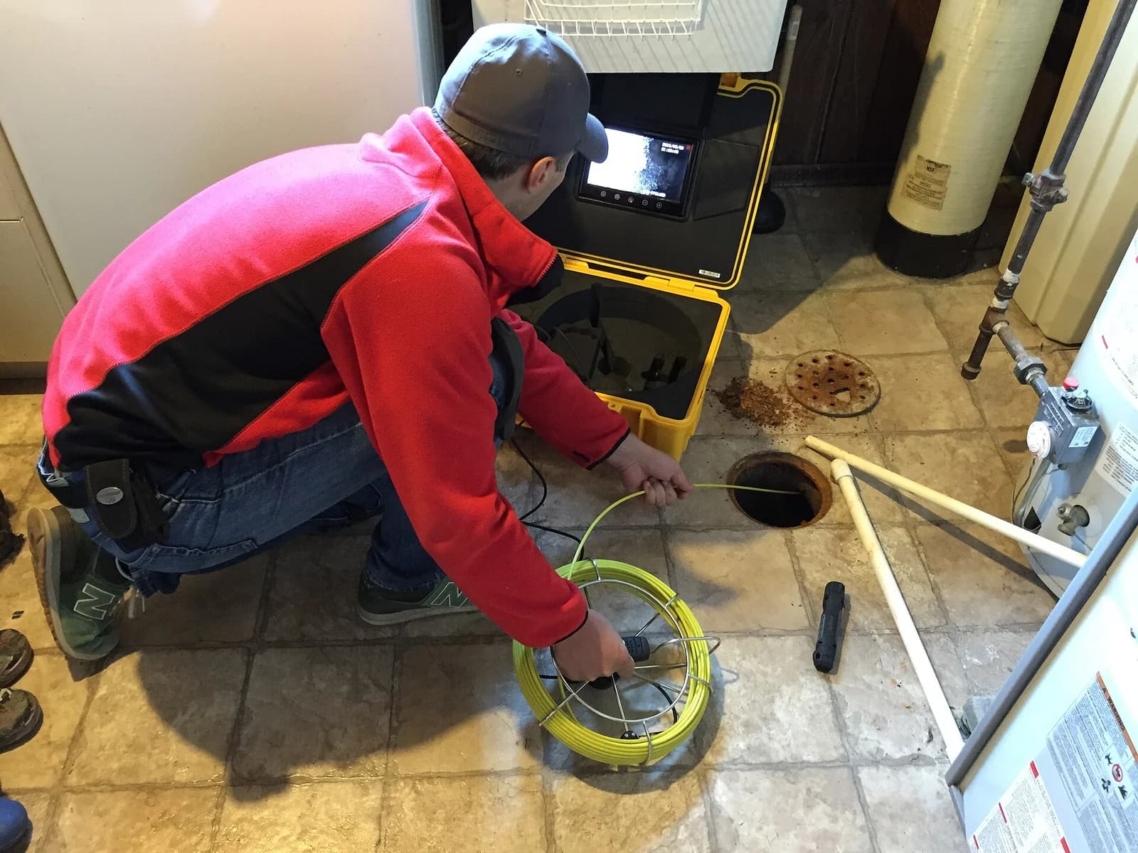 Plumber feeding a sewer inspection camera into a basement drain line with the monitor open beside him