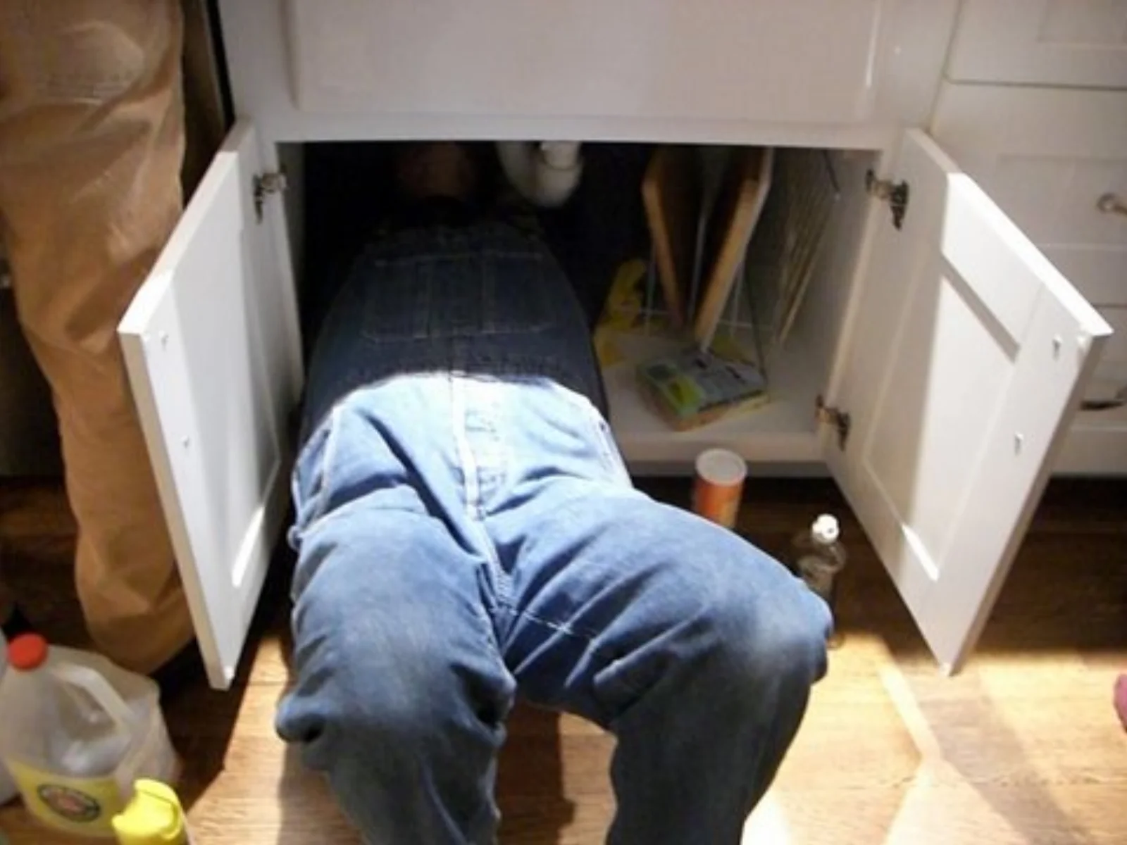 Plumber working inside an open kitchen sink cabinet on the supply and drain lines
