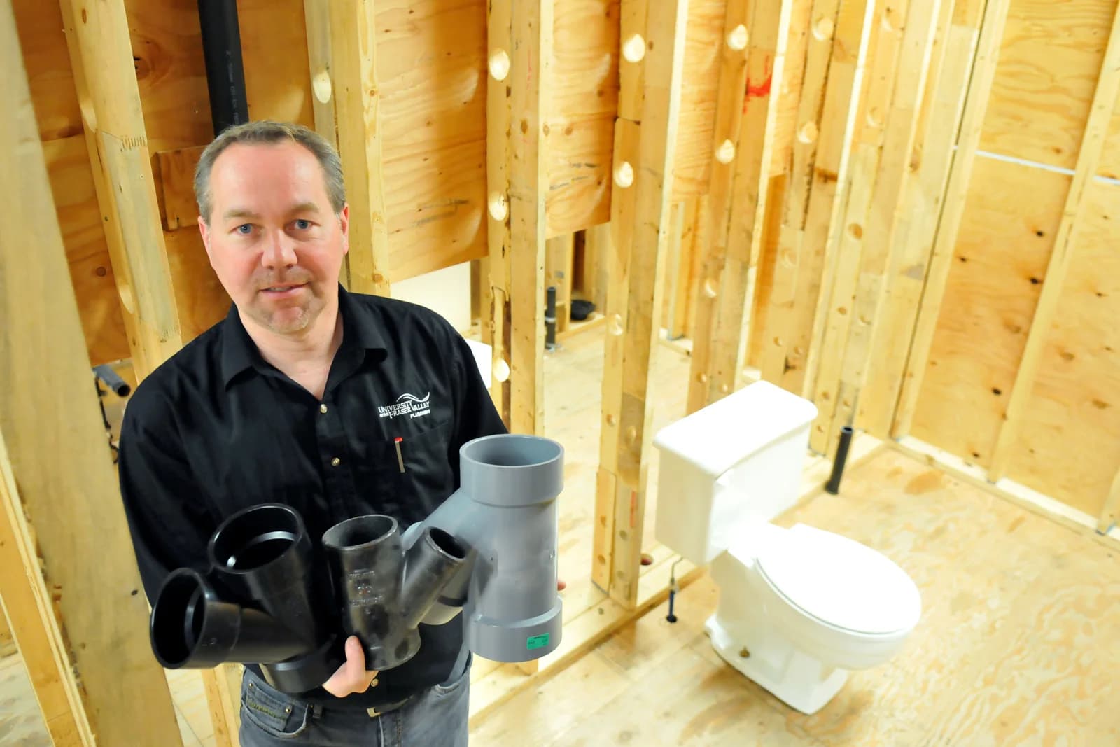Plumber holding DWV pipe fittings in a new-construction bathroom with a toilet and open wall studs